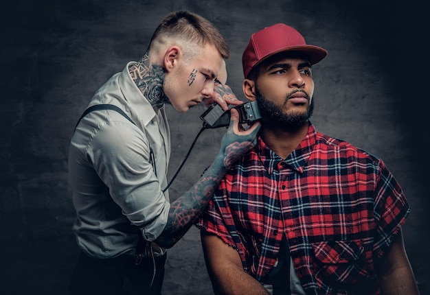 Un peluquero caucásico tatuado cortando la barba a un hombre negro con estilo.