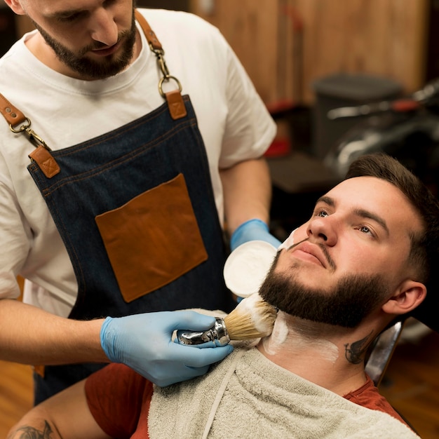 Foto gratuita peluquero con crema de afeitar para contornear la barba del cliente masculino