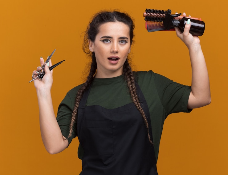 Foto gratuito peluquero femenino joven complacido en uniforme levantando herramientas de peluquero aisladas en la pared naranja
