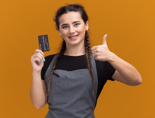 Peluquero femenino joven sonriente en uniforme que sostiene la tarjeta de crédito que muestra el pulgar hacia arriba aislado en la pared naranja