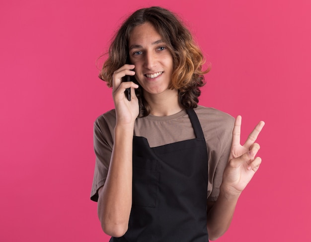 Foto gratuita peluquero guapo joven alegre con uniforme mirando al frente hablando por teléfono haciendo el signo de la paz aislado en la pared rosa con espacio de copia