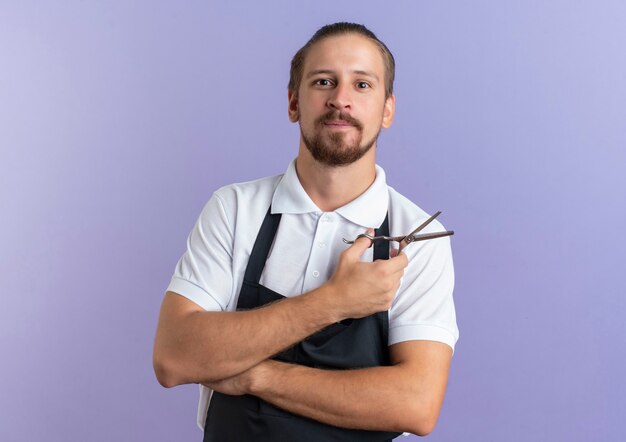 Peluquero guapo joven complacido vistiendo uniforme sosteniendo tijeras aisladas en la pared púrpura