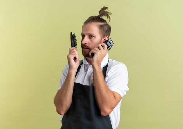 Peluquero guapo joven confiado con peine y cortapelos finge hablar por teléfono usando cortapelos como teléfono aislado en verde oliva con espacio de copia