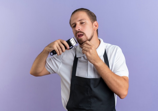 Peluquero guapo joven confiado con uniforme sosteniendo cortapelos y recortando su propia barba tocando su barbilla aislada en púrpura