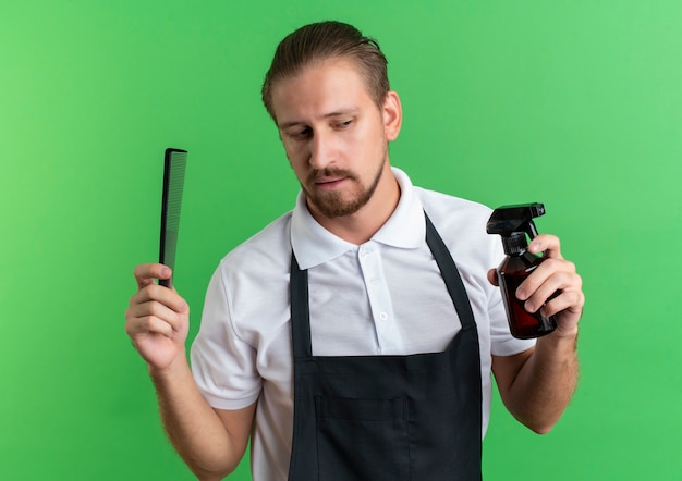 Peluquero guapo joven confiado con uniforme sosteniendo peine y botella de spray mirando al lado aislado en verde