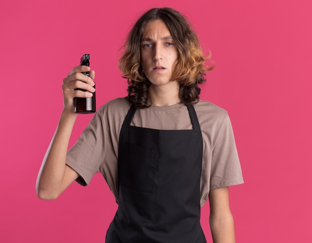 Peluquero guapo joven confiado vistiendo uniforme con botella de spray aislada en la pared rosa