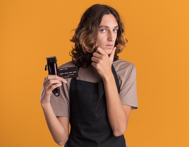 Peluquero guapo joven confiado vistiendo uniforme con tarjeta de crédito y cortapelos manteniendo la mano en la barbilla