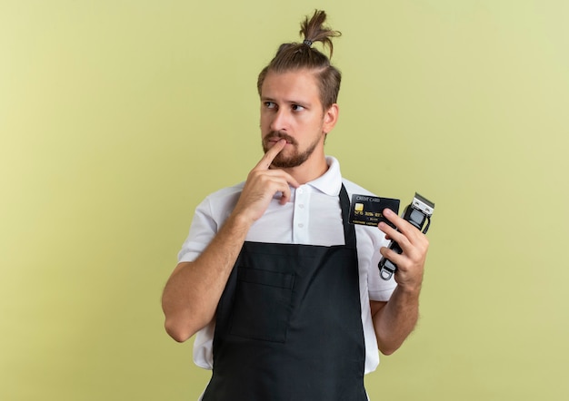 Foto gratuita peluquero guapo joven pensativo con tarjeta de crédito y cortapelos poniendo el dedo en el labio mirando al lado aislado en la pared verde oliva