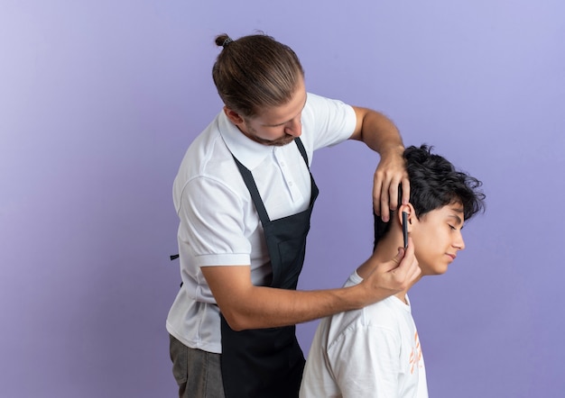 Peluquero guapo joven con uniforme de pie en la vista de perfil haciendo corte de pelo para cliente joven aislado sobre fondo púrpura con espacio de copia