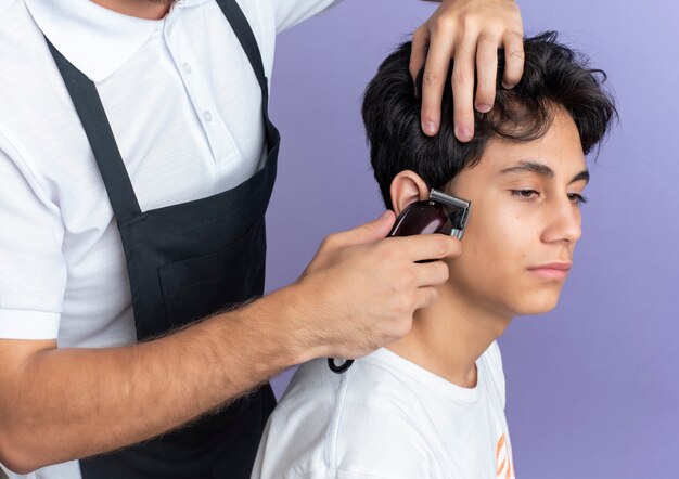 Peluquero guapo joven vistiendo uniforme haciendo corte de pelo para cliente joven aislado sobre fondo púrpura