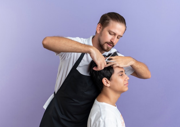 Peluquero guapo joven vistiendo uniforme haciendo corte de pelo para cliente joven aislado sobre fondo púrpura