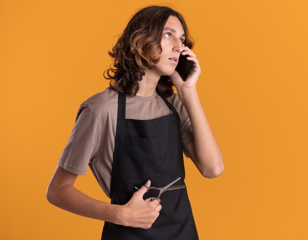 Peluquero guapo joven vistiendo uniforme sosteniendo tijeras hablando por teléfono mirando hacia arriba aislado en la pared naranja con espacio de copia