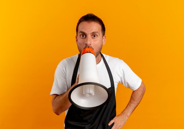 Peluquero hombre en delantal gritando por megáfono parado sobre pared naranja