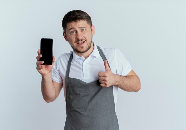 Peluquero hombre en delantal mostrando smartphone mostrando Thumbs up sonriendo confiados de pie sobre fondo blanco.