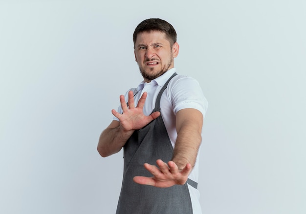 Peluquero hombre en delantal sosteniendo la mano haciendo gesto de defensa con expresión de disgusto de pie sobre fondo blanco.