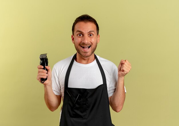 Peluquero hombre en delantal sosteniendo la máquina de corte de pelo apretando el puño feliz y emocionado de pie sobre la pared de olivo