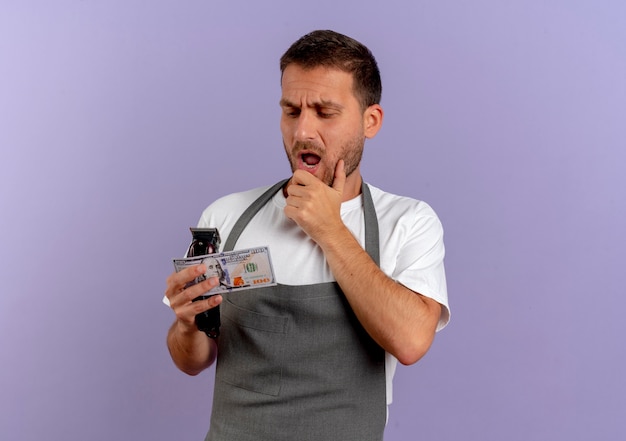 Peluquero hombre en delantal sosteniendo la máquina de corte de pelo y efectivo mirando asombrado y sorprendido de pie sobre la pared púrpura