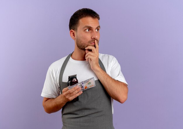 Peluquero hombre en delantal sosteniendo la máquina de corte de pelo y efectivo mirando a un lado pensando de pie sobre la pared púrpura