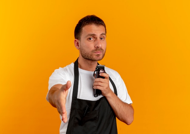 Peluquero hombre en delantal sosteniendo la máquina de corte de pelo mirando hacia el frente ofreciendo saludo de mano de pie sobre la pared naranja