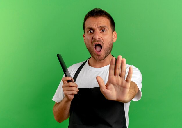 Peluquero hombre en delantal sosteniendo peine haciendo señal de pare con la mano gritando de pie sobre la pared verde