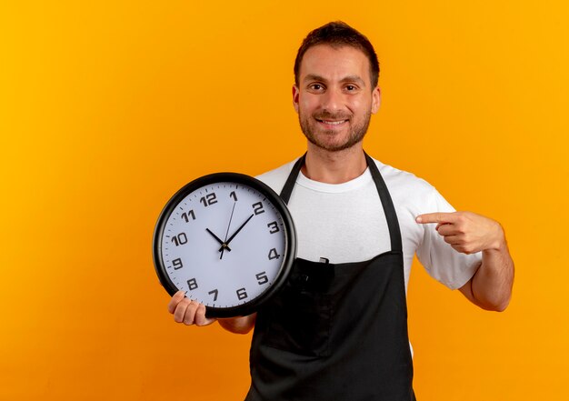 Peluquero hombre en delantal sosteniendo el reloj de pared apuntando con el dedo a él sonriendo confiado de pie sobre la pared naranja