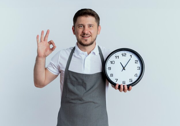 Peluquero hombre en delantal sosteniendo reloj de pared mostrando signo ok sonriendo de pie sobre fondo blanco.
