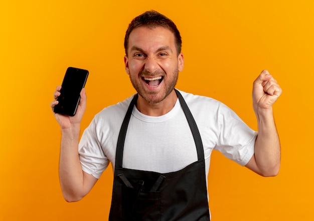 Peluquero hombre en delantal sosteniendo smartphone apretando el puño mirando hacia el frente feliz y emocionado de pie sobre la pared naranja