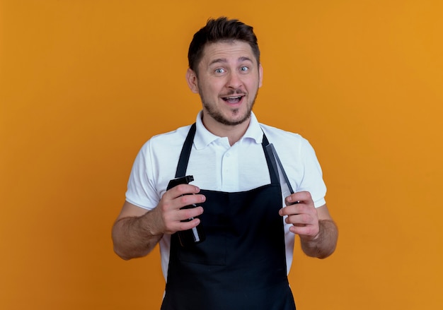 Peluquero hombre en delantal sosteniendo spray y tijeras mirando a la cámara sonriendo con cara feliz de pie sobre fondo naranja