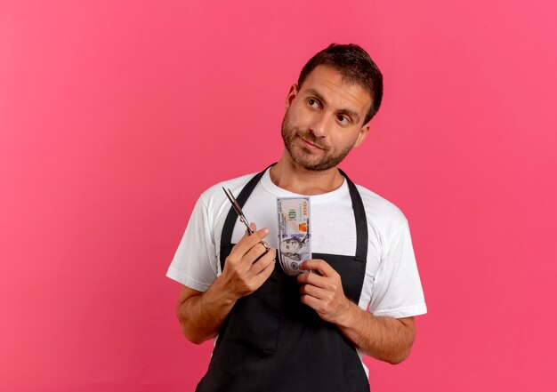 Peluquero hombre en delantal sosteniendo tijeras y efectivo mirando a un lado con expresión escéptica de pie sobre la pared rosa