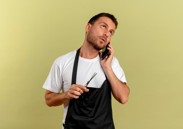 Peluquero hombre en delantal sosteniendo tijeras hablando por teléfono móvil con cara seria de pie sobre la pared de luz