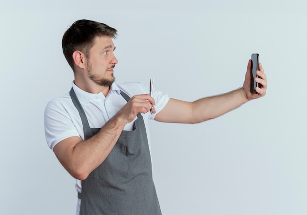 Peluquero hombre en delantal sosteniendo tijeras tomando una foto de sí mismo con smartphone parado sobre fondo blanco.