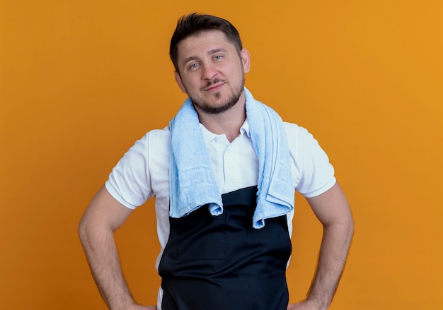 Foto gratuita peluquero hombre en delantal con una toalla alrededor del cuello lookign a la cámara sonriendo confiados de pie sobre fondo naranja