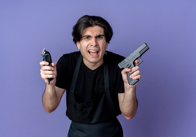 Peluquero hombre guapo joven enojado en uniforme sosteniendo cortapelos con pistola aislado sobre fondo azul.