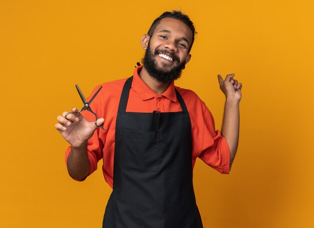 Peluquero masculino afroamericano joven alegre que lleva uniforme que sostiene las tijeras que apuntan detrás aislado en la pared anaranjada