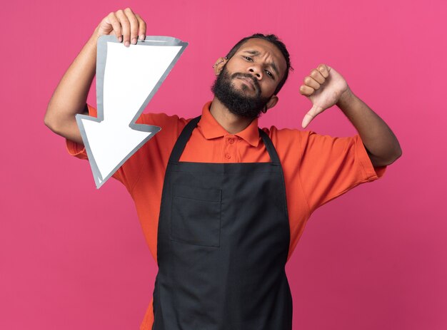 Peluquero masculino afroamericano joven disgustado con uniforme sosteniendo la marca de flecha que apunta hacia abajo mirando a la cámara mostrando el pulgar hacia abajo aislado sobre fondo rosa