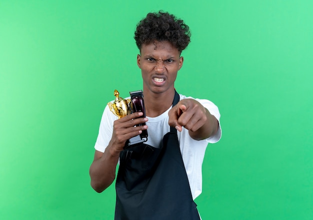 Peluquero masculino afroamericano joven enojado que lleva uniforme que sostiene el ganador de la copa y que le muestra gesto aislado en fondo verde