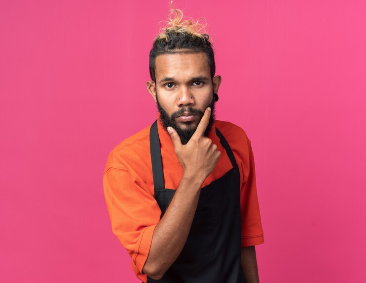 Foto gratuito peluquero masculino afroamericano joven pensativo vistiendo uniforme manteniendo la mano en la barbilla aislada en la pared rosa con espacio de copia