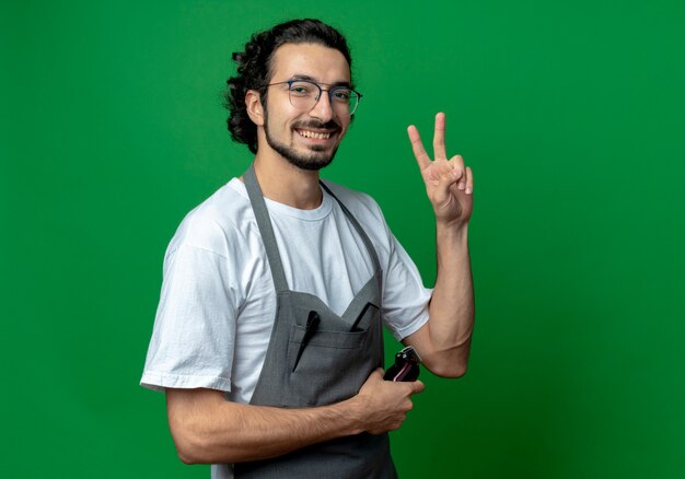 Foto gratuita peluquero masculino caucásico joven alegre con gafas y banda para el cabello ondulado en uniforme sosteniendo cortapelos y haciendo el signo de la paz aislado sobre fondo verde con espacio de copia