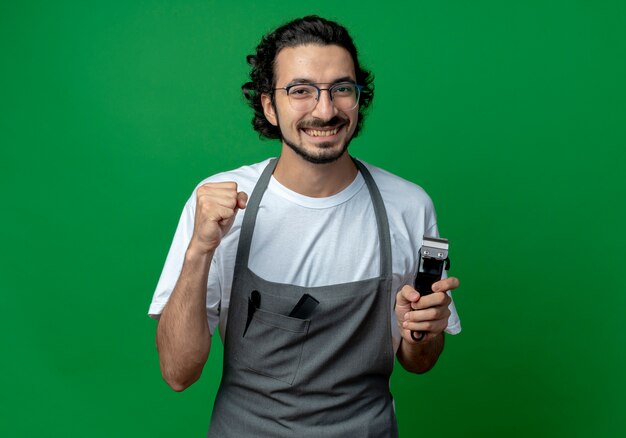 Peluquero masculino caucásico joven alegre con gafas y banda para el pelo ondulado en uniforme sosteniendo cortapelos y puño cerrado aislado sobre fondo verde con espacio de copia