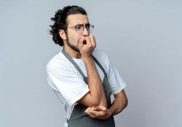 Peluquero masculino caucásico joven ansioso con gafas y banda para el pelo ondulado en uniforme poniendo la mano en la barbilla y otra en el codo mirando directamente aislado sobre fondo blanco con espacio de copia