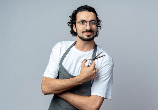 Peluquero masculino caucásico joven complacido con gafas y banda para el cabello ondulado en uniforme de pie con postura cerrada y sosteniendo tijeras aisladas sobre fondo blanco con espacio de copia