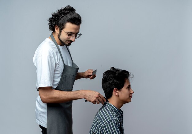 Peluquero masculino caucásico joven complacido con gafas y banda para el cabello ondulado en uniforme de pie en vista de perfil haciendo corte de pelo para su joven cliente aislado sobre fondo blanco con espacio de copia