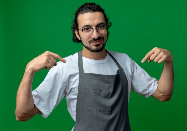 Foto gratuita peluquero masculino caucásico joven confiado con uniforme y gafas apuntando a sí mismo aislado sobre fondo verde