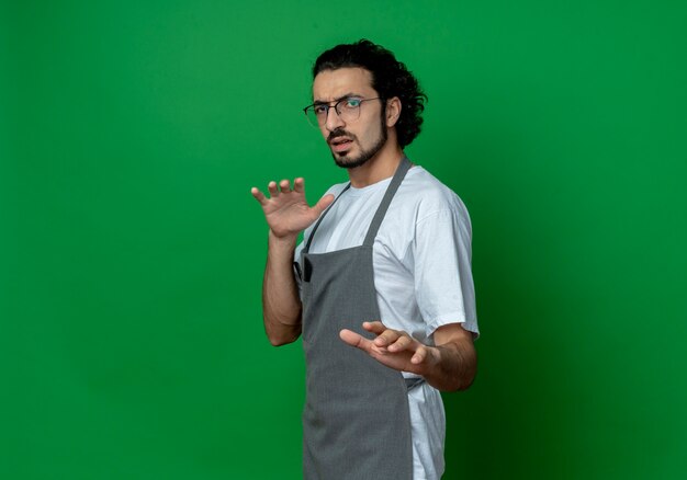 Peluquero masculino caucásico joven descontento con gafas y banda para el pelo ondulado en uniforme estirando la mano a la cámara gesticulando no aislado sobre fondo verde con espacio de copia