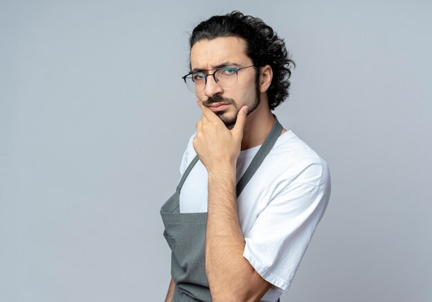 Peluquero masculino caucásico joven pensativo con gafas y banda para el pelo ondulado en uniforme de pie en vista de perfil tocando la barbilla aislada sobre fondo blanco con espacio de copia