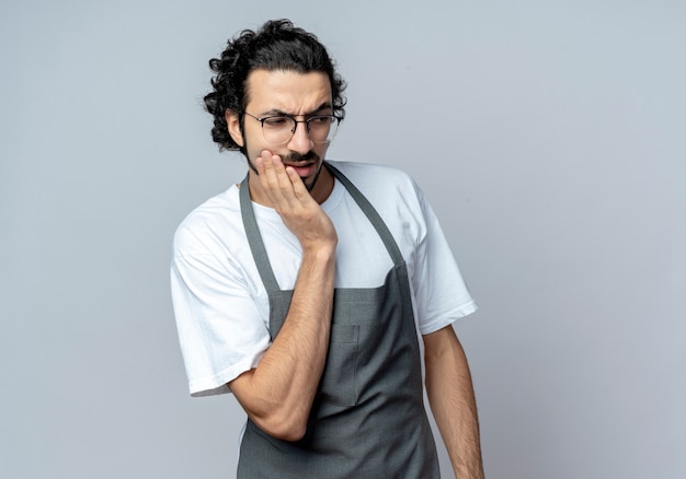 Peluquero masculino caucásico joven pensativo con gafas y banda para el pelo ondulado en uniforme poniendo la mano en la cara mirando al lado aislado sobre fondo blanco con espacio de copia