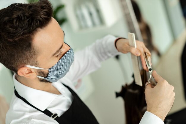 Peluquero masculino cortando el cabello del cliente mientras usa mascarilla durante la epidemia de coronavirus