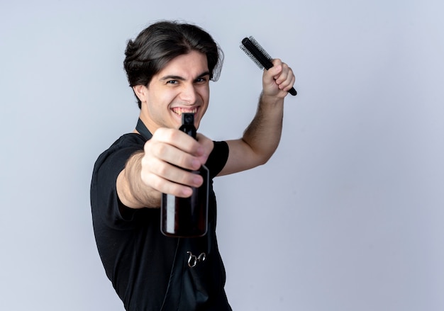 Peluquero masculino guapo joven alegre en uniforme sosteniendo la botella de spray en la cámara y levantando el peine aislado sobre fondo blanco