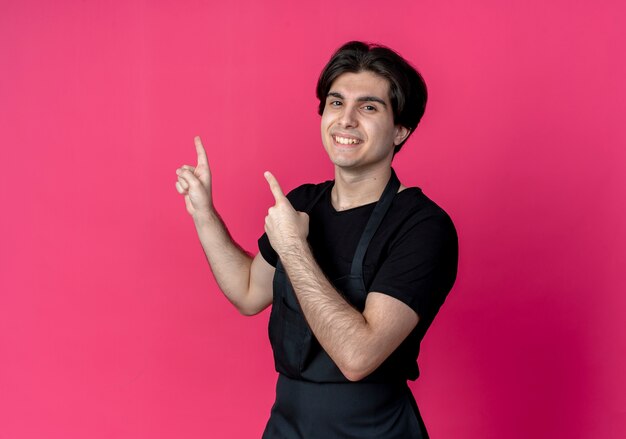 Peluquero masculino guapo joven sonriente en puntos uniformes en el lado aislado en rosa con espacio de copia