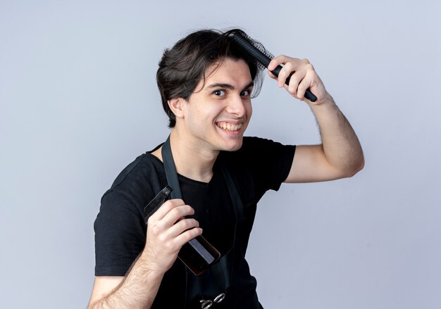 Peluquero masculino guapo joven sonriente en uniforme peinándose y sosteniendo una botella de spray aislado en blanco